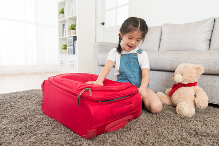 happy little girl closing luggage suitcase in living room when she finishing packing and ready to travel with teddy bear